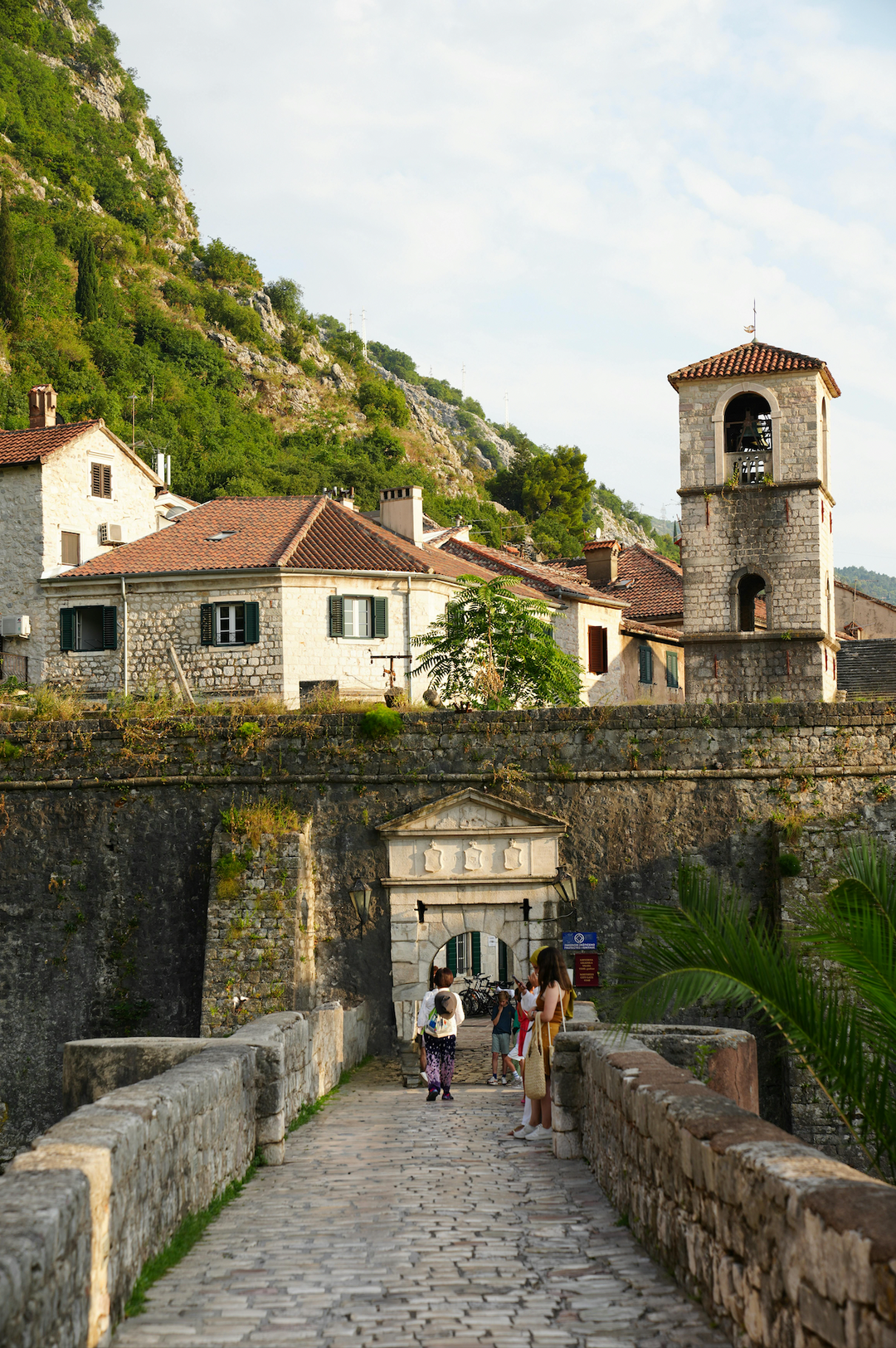 Stone bridge leading to an old town gate below a bell tower and hills