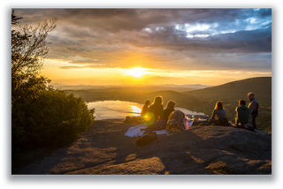 Group of friends sitting on a hilltop watching the sunset over a lake