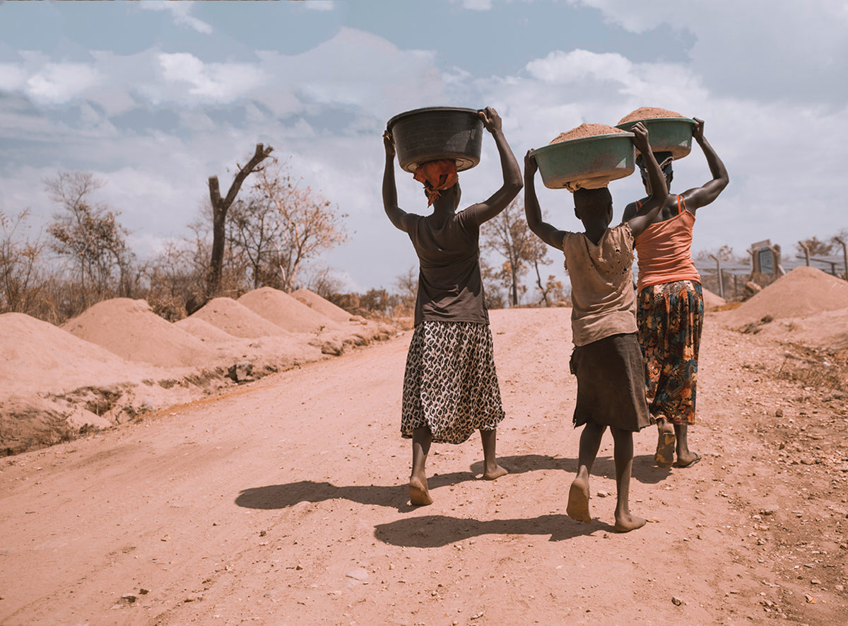 Three women carrying bowls on their heads along a red dirt road