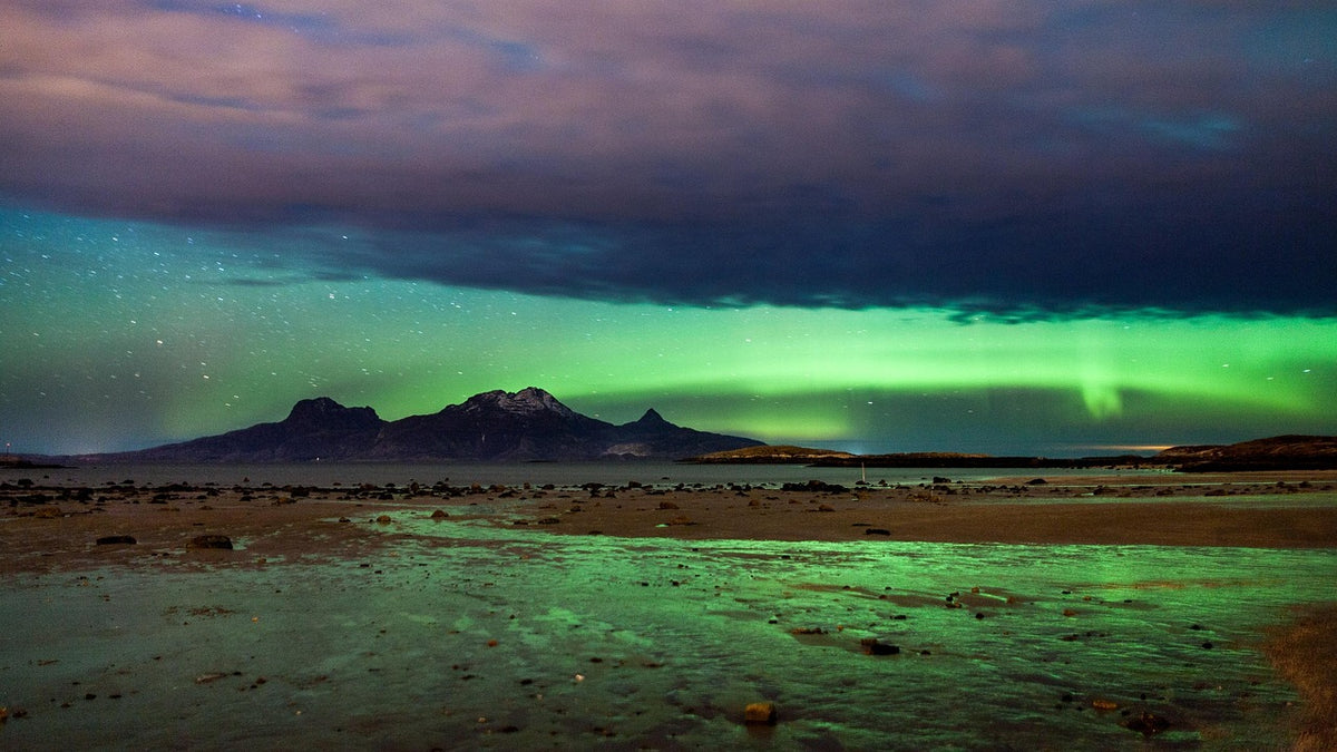Green northern lights over a wet shoreline and distant mountains